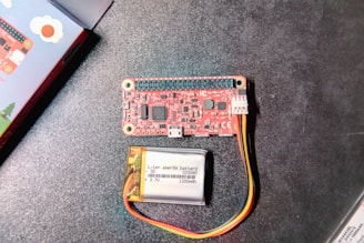 A red electronic circuit board with multiple components, connectors, and labeled parts alongside a small lithium polymer battery with yellow, red, and black wires. The background is a dark tabletop surface. A colorful box is partially visible on the left edge.