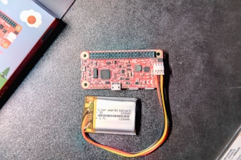 A red electronic circuit board with multiple components, connectors, and labeled parts alongside a small lithium polymer battery with yellow, red, and black wires. The background is a dark tabletop surface. A colorful box is partially visible on the left edge.