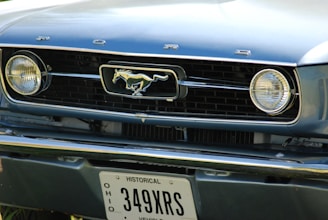 A close-up of the front grille of a classic Ford Mustang, featuring the iconic horse emblem in the center with 'Ford' lettering above. The headlights are round and prominent, and a historical Ohio license plate is visible at the bottom.
