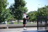 A man is playing the bagpipes outdoors in a park setting, surrounded by lush green trees. He is dressed in a traditional kilt and white shirt with a black hat and long socks. The background has a paved area with grass, trees, and some distant people visible.
