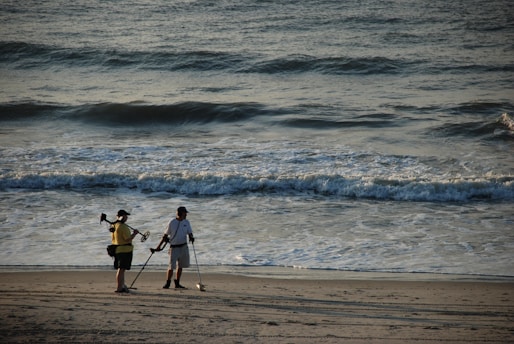 Two people are standing on the beach near the shoreline, each holding a metal detector. The ocean is in the background with waves gently rolling in. The scene suggests a quiet and focused search for metal objects or treasures in the sand.