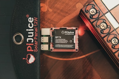 A PiJuice solar panel is shown alongside its battery unit, specifically a BP7X model, and two AA batteries. The background features a wooden surface with a decorative box partially visible, displaying numbers on its side.