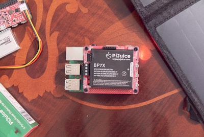 A PiJuice Battery Module with labeled ports and connections is placed on a wooden surface. Adjacent are several cables, a small circuit board, and part of a laptop or tablet case.