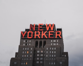 A tall building with a prominent red neon sign that spells 'NEW YORKER' atop its structure. The building features multiple windows and is set against a cloudy sky.