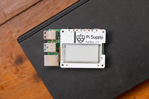 A small electronic board labeled 'Pi Supply PaPiRus v1.9' featuring an e-paper display is situated on a closed black book. The board includes several USB ports and is placed on a wooden surface.