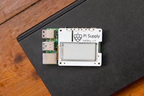 A small electronic board labeled 'Pi Supply PaPiRus v1.9' featuring an e-paper display is situated on a closed black book. The board includes several USB ports and is placed on a wooden surface.
