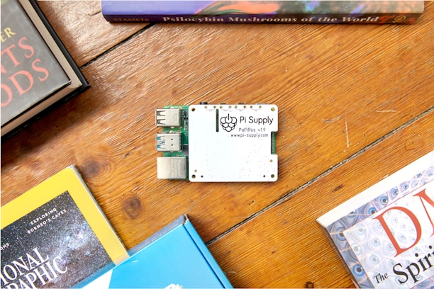 A Pi Supply electronic board is placed on a wooden surface surrounded by books. The board features USB ports and is labeled with its brand and model. The books surrounding it have colorful covers and appear to be related to science and exploration.