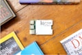 A Pi Supply electronic board is placed on a wooden surface surrounded by books. The board features USB ports and is labeled with its brand and model. The books surrounding it have colorful covers and appear to be related to science and exploration.