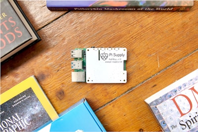 A Pi Supply electronic board is placed on a wooden surface surrounded by books. The board features USB ports and is labeled with its brand and model. The books surrounding it have colorful covers and appear to be related to science and exploration.