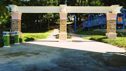 A landscaped area features a decorative archway with stone pillars and pastel-colored accents. Beyond the archway, a children's playground with blue climbing structures and slides is visible. The surroundings are lush with greenery, including neatly trimmed grass and trees providing shade.