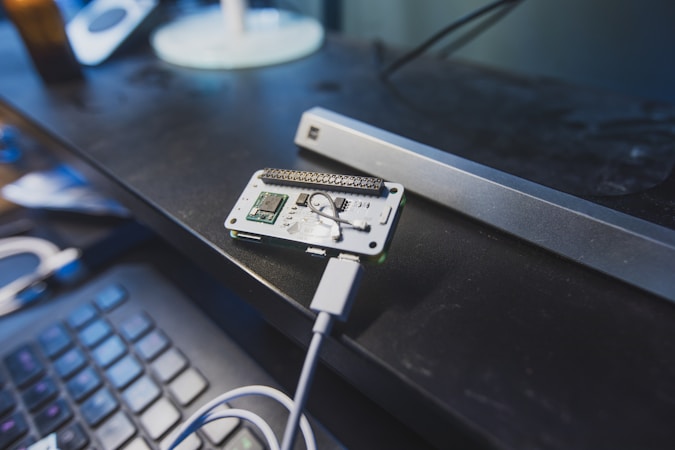 A small electronic circuit board is placed on a black desk, connected to a device via a cable. Nearby, there is a keyboard with visible keys. The workspace has a tech-focused environment with various gadgets and components.
