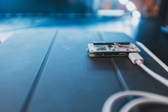 A small circuit board lies on a dark surface, connected to a white USB cable. The surrounding lighting creates a soft, focused atmosphere, with a shallow depth of field emphasizing the electronic components.
