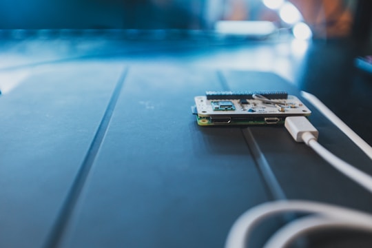 A small circuit board lies on a dark surface, connected to a white USB cable. The surrounding lighting creates a soft, focused atmosphere, with a shallow depth of field emphasizing the electronic components.