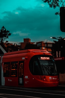 A red tram with illuminated destination sign 'Newcastle Beach' is stationed on a street. The tram is surrounded by urban buildings in the background, under a moody, cloudy sky. The scene is captured during the evening or late afternoon, with streetlights adding a warm glow.