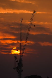 A hydraulic crane lifting heavy equipment against a sunset sky with black and yellow tones.