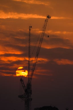 A hydraulic crane lifting heavy equipment against a sunset sky with black and yellow tones.