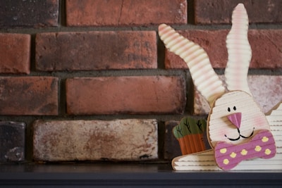 A whimsical cardboard cutout of a bunny with large corrugated ears and a pink bow tie is placed against a background of red and brown brick. The bunny has simple facial features and is accompanied by a small cardboard pot with carrots on its left.