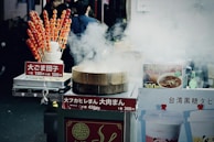 A street food stall is surrounded by steam, with a bamboo steamer prominently in front. The stall features various signs with pricing and food names written in Japanese. On the left, skewers of candied fruit or dango are displayed in a holder. To the right, there are images of soup and bubble tea on display. A few beverages, including a green bottle, can be seen among the food items.