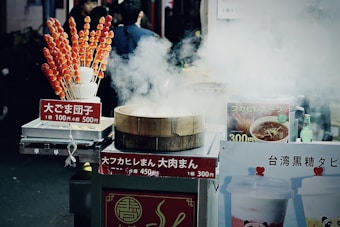 A street food stall is surrounded by steam, with a bamboo steamer prominently in front. The stall features various signs with pricing and food names written in Japanese. On the left, skewers of candied fruit or dango are displayed in a holder. To the right, there are images of soup and bubble tea on display. A few beverages, including a green bottle, can be seen among the food items.