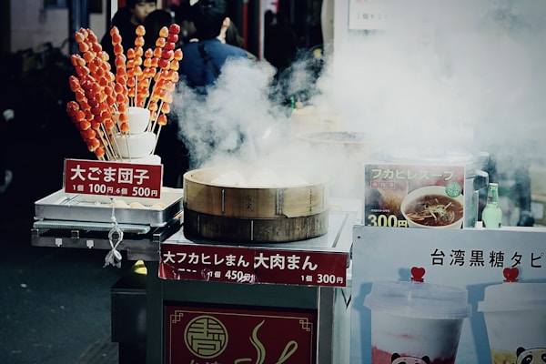 A street food stall is surrounded by steam, with a bamboo steamer prominently in front. The stall features various signs with pricing and food names written in Japanese. On the left, skewers of candied fruit or dango are displayed in a holder. To the right, there are images of soup and bubble tea on display. A few beverages, including a green bottle, can be seen among the food items.