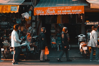 People are standing in line in front of a small market shop with signs in Vietnamese. There are several scooters parked along the street, and the shop is covered with some merchandise, including colorful plastic containers. The scene appears to be in an urban setting with a casual atmosphere.