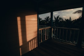 A wooden porch with railings overlooks a view filled with lush palm trees. The warm glow of the setting sun casts long shadows on the wooden floor and wall, creating an inviting and peaceful atmosphere.