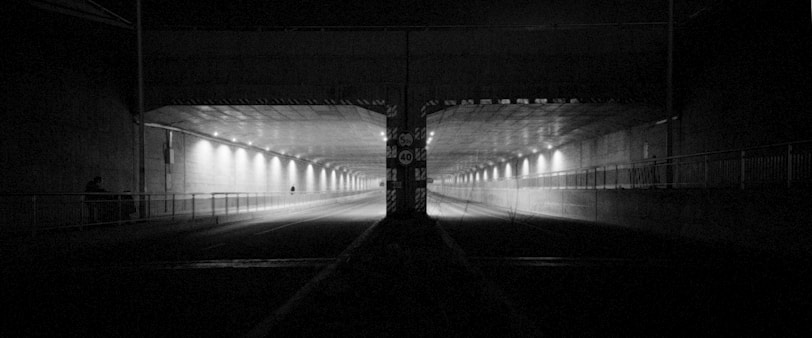 A dimly lit tunnel with symmetrical lighting on both sides. The tunnel walls are lined with evenly spaced lamps that create a repetitive pattern of light and shadow. The scene is devoid of traffic, adding to the empty and desolate feel. The central column divides the tunnel into two similar pathways.