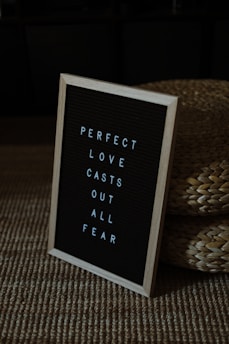 A framed message board with white letters on a black background reads 'PERFECT LOVE CASTS OUT ALL FEAR'. It is placed on a textured woven mat, next to straw baskets. The setting appears cozy and somewhat dimly lit.