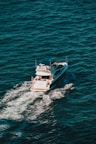 A white yacht cruises through deep blue ocean waters, creating a trail of white foam behind it. The vessel has a sleek design with a visible deck where people can be seen relaxing.