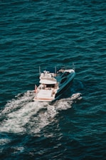 A white yacht cruises through deep blue ocean waters, creating a trail of white foam behind it. The vessel has a sleek design with a visible deck where people can be seen relaxing.
