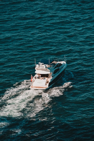 A white yacht cruises through deep blue ocean waters, creating a trail of white foam behind it. The vessel has a sleek design with a visible deck where people can be seen relaxing.