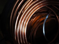 A close-up view of coiled copper wires, showcasing their shiny and smooth surface texture. The coils overlap each other, creating an intricate pattern with reflections and shadows that add depth to the image.