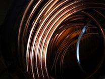 A close-up view of coiled copper wires, showcasing their shiny and smooth surface texture. The coils overlap each other, creating an intricate pattern with reflections and shadows that add depth to the image.