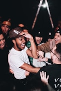 A group of people, mostly young men, are gathered closely together, laughing and engaging in lively interaction. They appear to be enjoying themselves in a social setting, with the focal point being a person in a white shirt and baseball cap. The mood is energetic and cheerful, with hands reaching out toward each other.