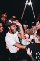 A group of people, mostly young men, are gathered closely together, laughing and engaging in lively interaction. They appear to be enjoying themselves in a social setting, with the focal point being a person in a white shirt and baseball cap. The mood is energetic and cheerful, with hands reaching out toward each other.