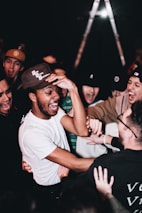 A group of people, mostly young men, are gathered closely together, laughing and engaging in lively interaction. They appear to be enjoying themselves in a social setting, with the focal point being a person in a white shirt and baseball cap. The mood is energetic and cheerful, with hands reaching out toward each other.
