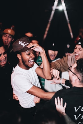 A group of people, mostly young men, are gathered closely together, laughing and engaging in lively interaction. They appear to be enjoying themselves in a social setting, with the focal point being a person in a white shirt and baseball cap. The mood is energetic and cheerful, with hands reaching out toward each other.