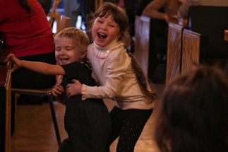 Two young children are joyfully playing together indoors. The older girl, with long hair and wearing a light-colored sweater, is smiling broadly as she holds a younger boy, who is in dark clothing, with one arm around his back. They appear to be having a fun time, as the boy leans forward with a playful expression. Surrounding them are other people seated at tables, suggesting a busy and warm gathering environment.