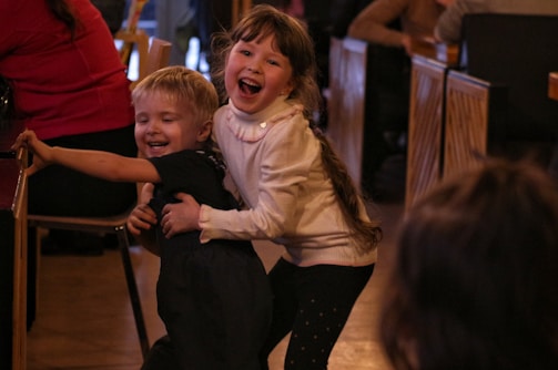 Two young children are joyfully playing together indoors. The older girl, with long hair and wearing a light-colored sweater, is smiling broadly as she holds a younger boy, who is in dark clothing, with one arm around his back. They appear to be having a fun time, as the boy leans forward with a playful expression. Surrounding them are other people seated at tables, suggesting a busy and warm gathering environment.