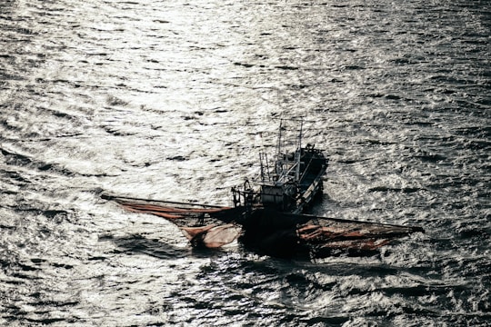 A fishing boat with large nets cast out on the sides floats on the water, with shimmering sunlight reflecting off the rippling surface of the sea.