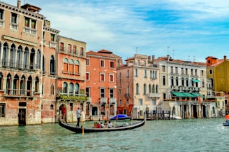 A picturesque canal scene featuring a gondola with passengers gliding through the water, surrounded by historic Venetian buildings. The architecture showcases a blend of ornate windows and vibrant colors, reflecting the charm and elegance of Venice.