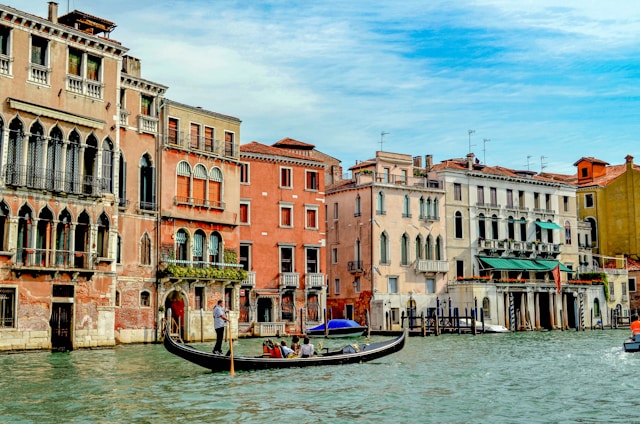 A picturesque canal scene featuring a gondola with passengers gliding through the water, surrounded by historic Venetian buildings. The architecture showcases a blend of ornate windows and vibrant colors, reflecting the charm and elegance of Venice.