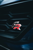 A close-up view of a car grille featuring a sleek and shiny emblem with the letters 'R' in red. The grille has a geometric pattern and is set against a dark background, creating a striking contrast.