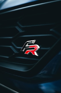 A close-up view of a car grille featuring a sleek and shiny emblem with the letters 'R' in red. The grille has a geometric pattern and is set against a dark background, creating a striking contrast.
