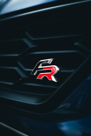 A close-up view of a car grille featuring a sleek and shiny emblem with the letters 'R' in red. The grille has a geometric pattern and is set against a dark background, creating a striking contrast.