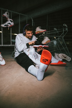 A person is sitting on the ground, wearing white socks and holding a sneaker with an orange sole. Multiple sneakers are scattered around, hanging from a metal fence and placed in a shopping cart. The person is wearing headphones and seems to be engaged with one of the sneakers, possibly lacing or examining it closely.