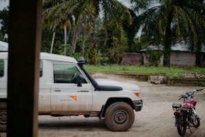 A white Land Cruiser with the World Vision logo is parked on a dirt road. A red motorcycle is nearby, with tropical trees and a simple building in the background, suggesting a rural setting.