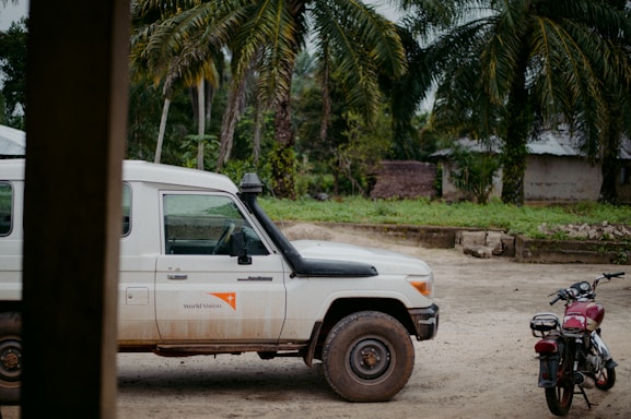 A white Land Cruiser with the World Vision logo is parked on a dirt road. A red motorcycle is nearby, with tropical trees and a simple building in the background, suggesting a rural setting.