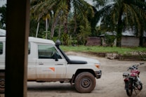 A white Land Cruiser with the World Vision logo is parked on a dirt road. A red motorcycle is nearby, with tropical trees and a simple building in the background, suggesting a rural setting.
