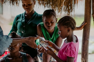A group of young individuals are gathered under a thatched roof, with a girl in a pink shirt attentively looking at photos. Another young girl in a green shirt, along with other children, is observing the scene closely. An adult's hand is reaching out, possibly passing the photos to them. The setting appears relaxed and informal.